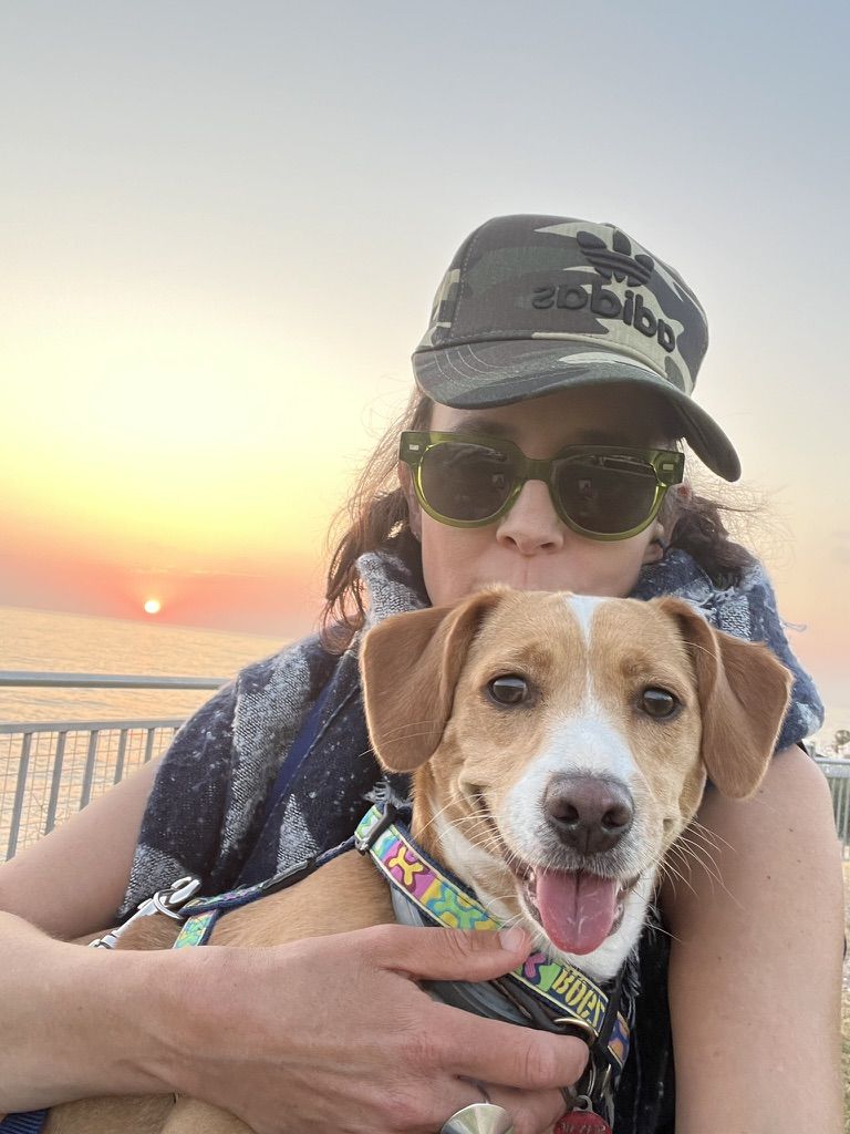 Hilary hugging a happy tan and white dog in front of a sunset over the sea